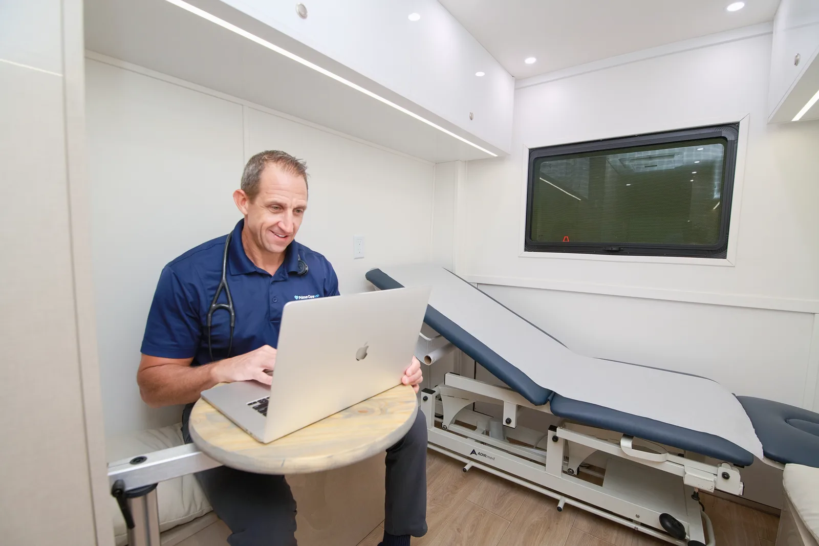 A PrimeCare provider working at a laptop beside the exam table inside the Mobile Medical Office